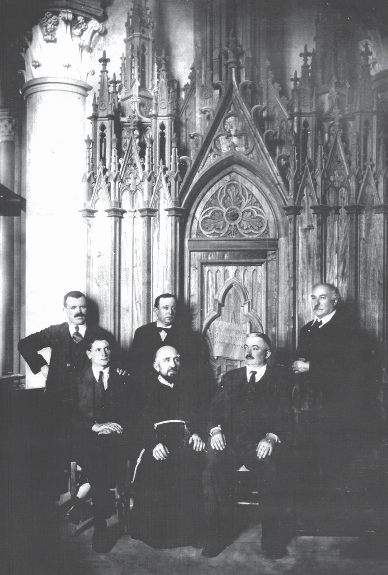 Retrato de grupo de seis homens posados diante de um altar de madeira entalhada em estilo gótico: três em pé e três sentados, um deles em hábito religioso.