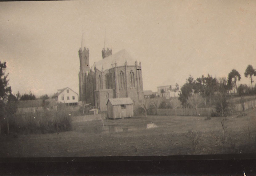 Vista distante da Catedral, com casas de madeira, cercas e vegetação em primeiro plano.