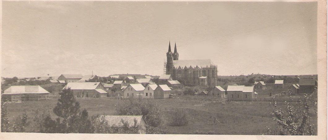 Vista distante de Vacaria com a Catedral ao fundo, as torres ainda cercadas por andaimes, e casas de madeira espalhadas pelo campo.