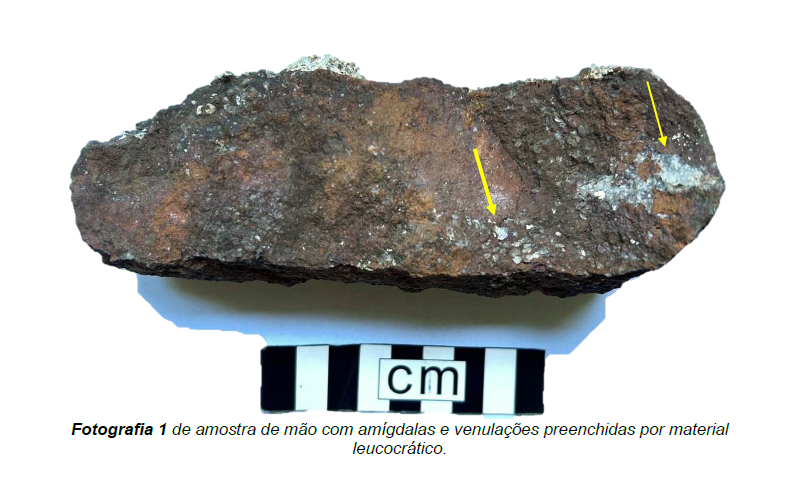 Fotografia da amostra de rocha analisada, com setas indicando as amígdalas preenchidas na superfície.